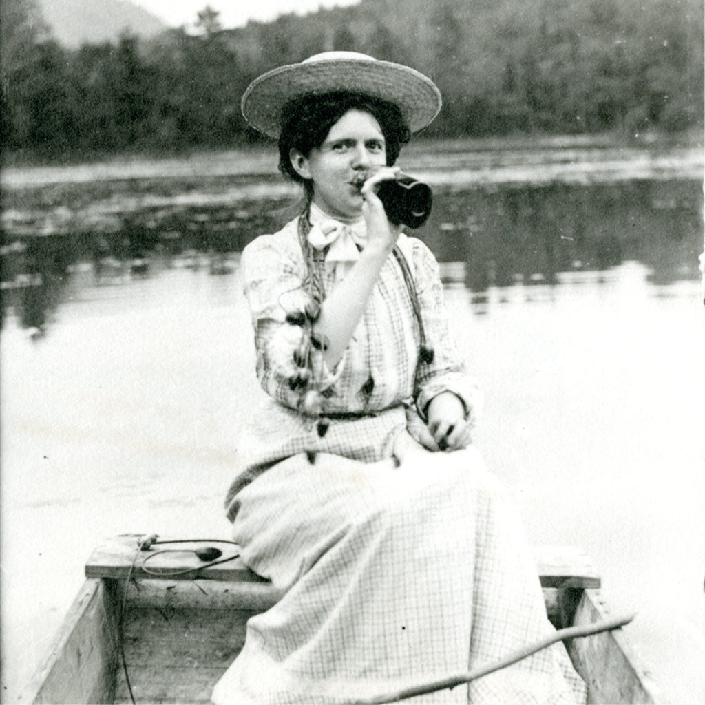 A black and white photograph of a woman wearing a wide-brimmed straw hat and long-sleeve full-length dress sitting on the back of a small row boat drinking from a dark colored glass bottle.