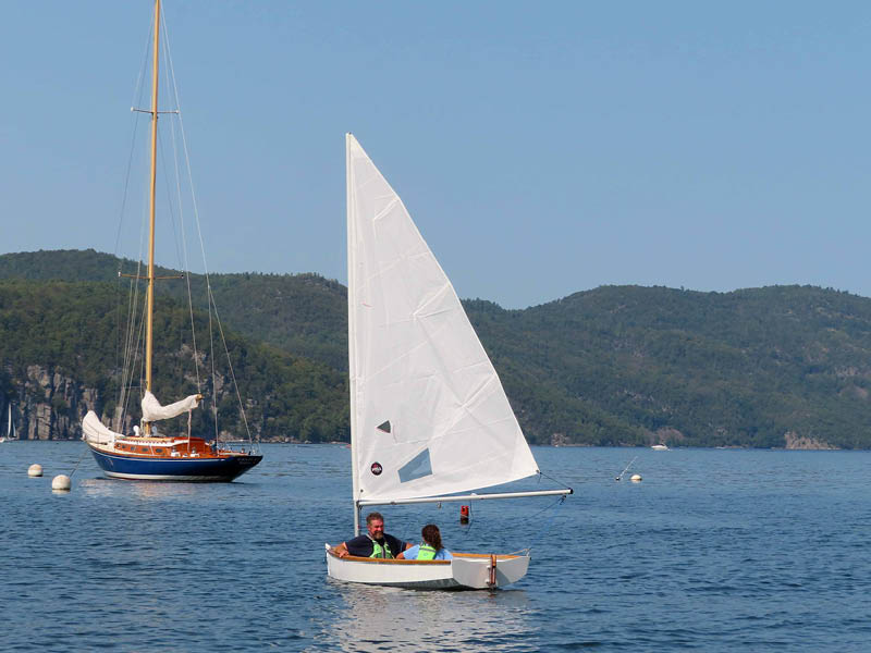 An adult and child in a small sailboat in front of a larger sailboat and mountains on a sunny day.