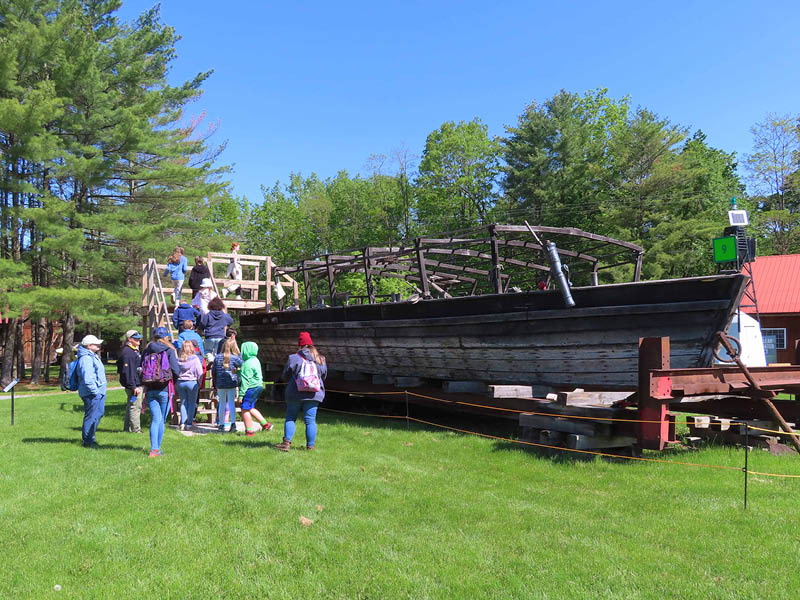 Replica wooden boat Philadelphia II sitting on a green lawn with blue skies and a group of children climbing aboard