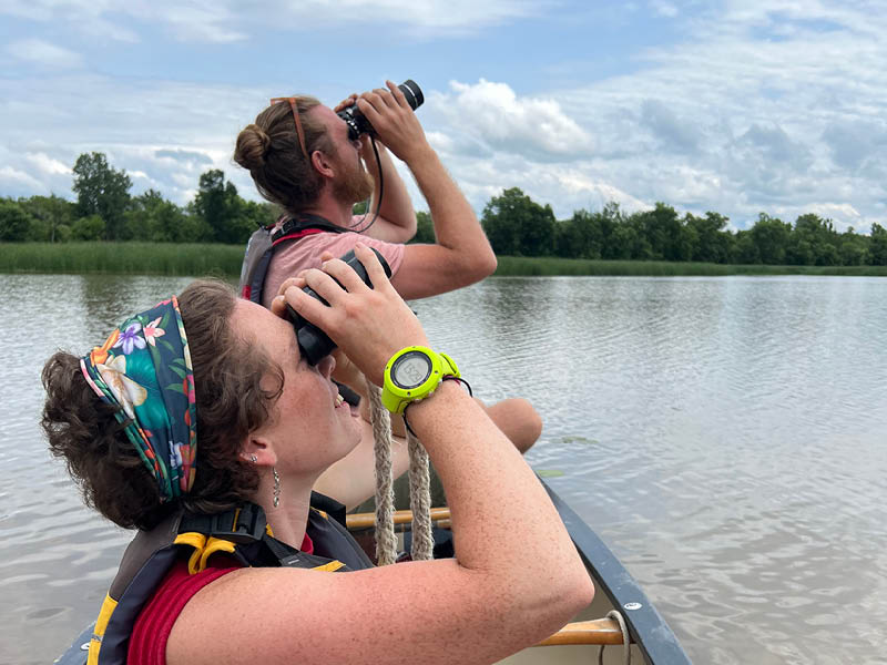 Two people sitting in a canoe and looking at the sky with binoculars.