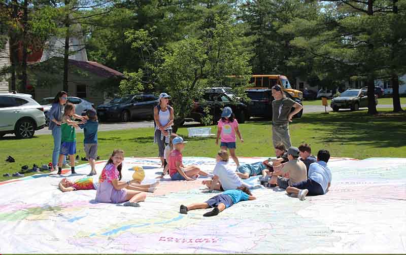 Several children and adults standing or lying on a very large white map outside.